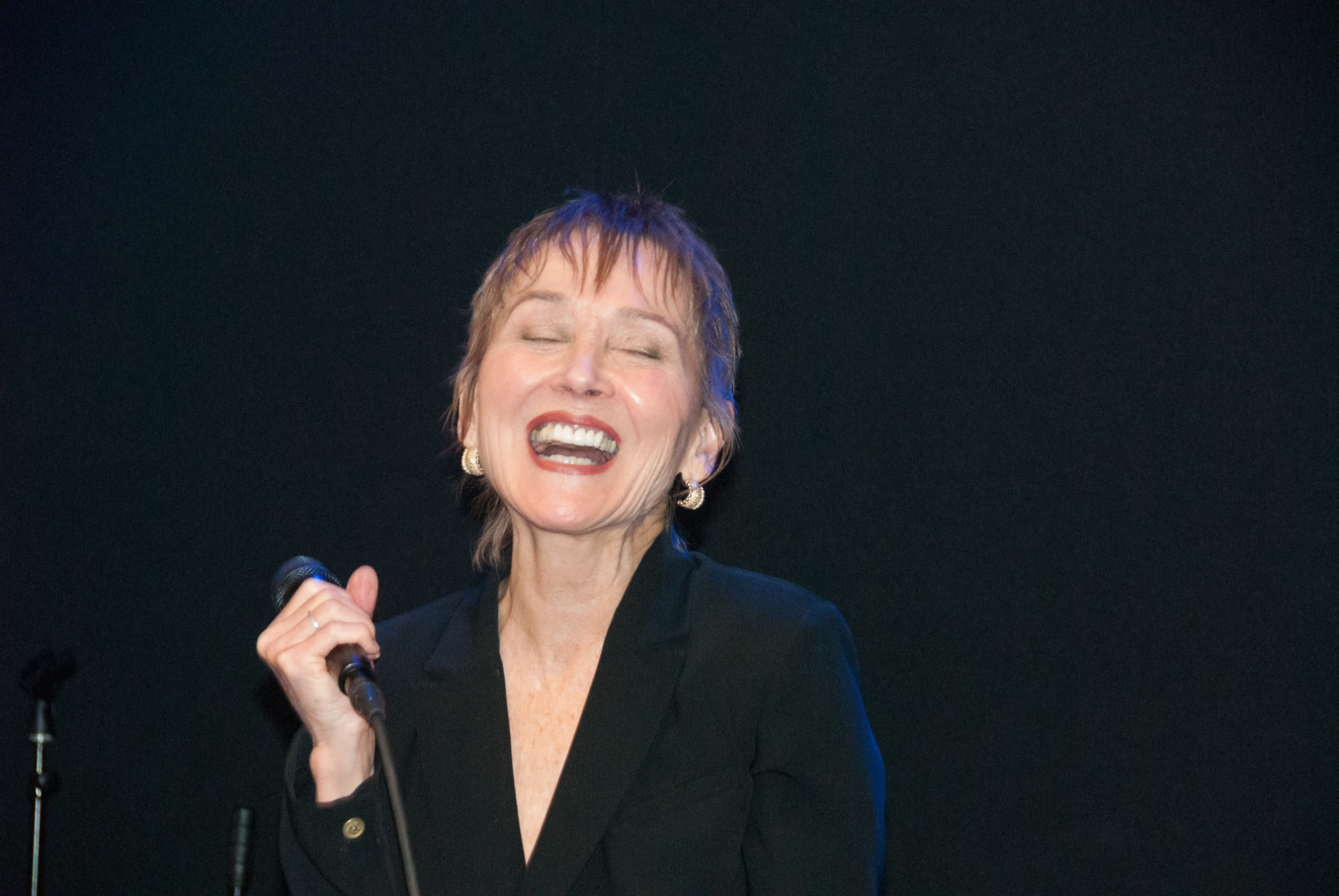 Audrey Moira Shimkas singing joyfully into a handheld microphone on a dark stage